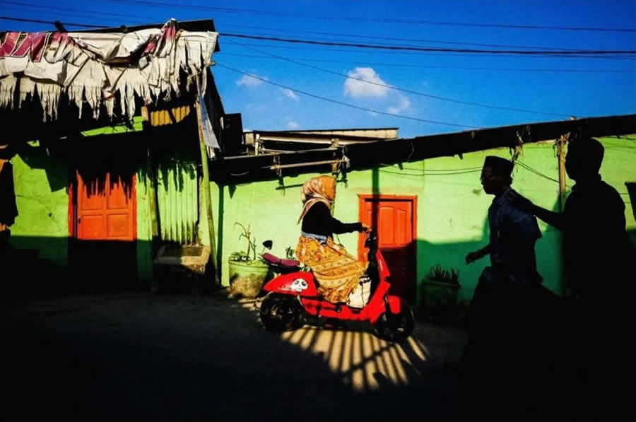 Woman riding a red scooter past a green wall with strong sunlight and shadows, while silhouettes of pedestrians create layered composition in a vibrant street photography scene.
