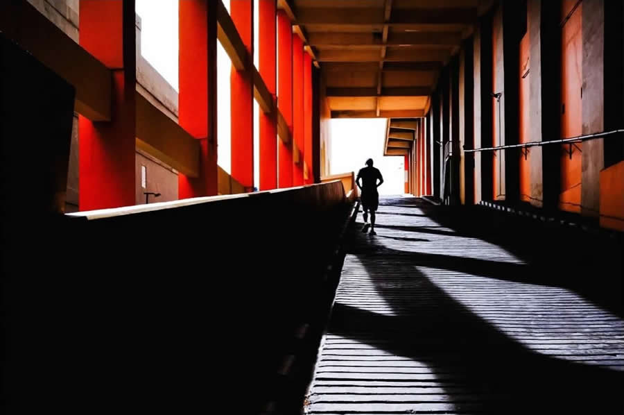 Silhouette of a person walking through a corridor with bold red columns, strong shadows, and leading lines, creating a dramatic geometric composition in street photography.