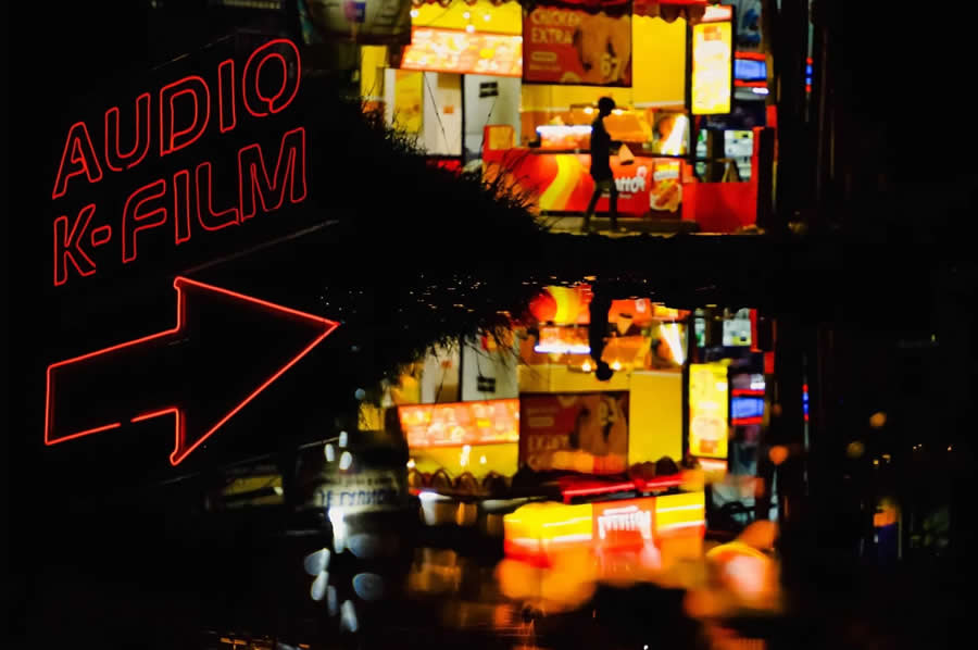 Neon sign reading &ldquo;Audio K-Film&rdquo; with bright street lights and a person walking, reflected in a wet surface, creating colorful reflections and a dynamic night street photography scene.