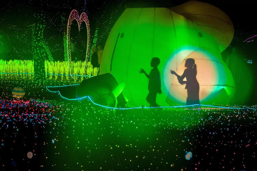 Silhouettes of two people interacting in front of bright green illuminated installation with colorful lights and reflections, creating a dreamy and surreal night street photography scene.