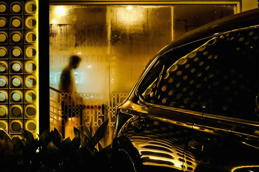 Blurred silhouette of a person walking behind a fogged glass window with warm golden lighting, alongside reflections on a parked car, creating a layered and atmospheric street photography composition.