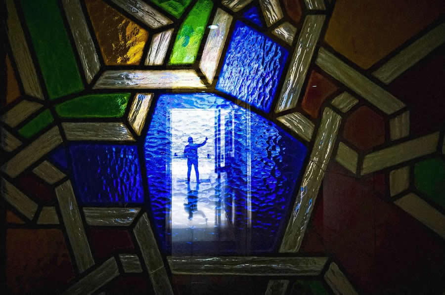 Silhouette of a person seen through a colorful stained glass frame with dominant blue light, creating a layered composition with geometric shapes and vibrant tones in street photography.