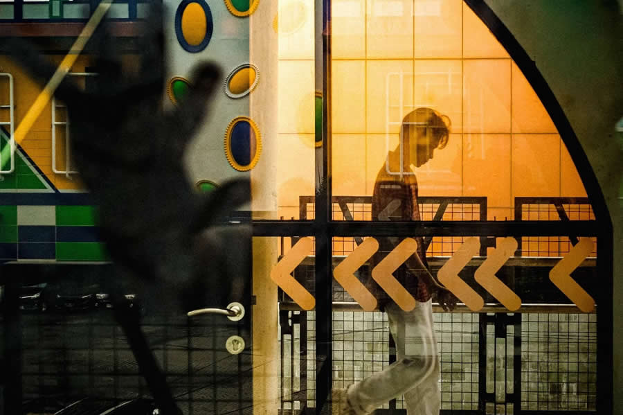 Person walking behind glass with warm orange lighting, layered with reflections and blurred foreground elements, featuring directional arrow patterns that create depth and motion in a street photography composition.