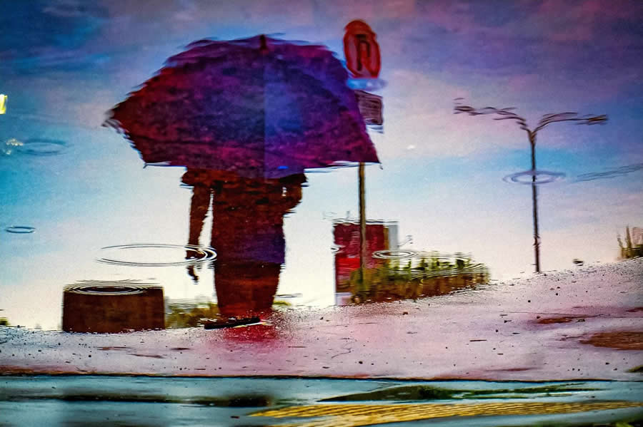 Reflection of a person holding an umbrella in a rain puddle, with ripples and vibrant colors creating a painterly and abstract street photography composition.