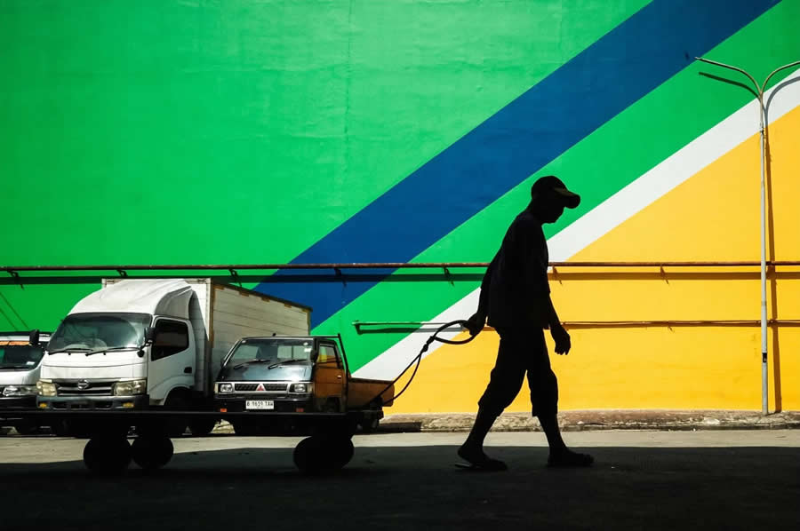 Silhouette of a person pulling a cart in front of a colorful wall with green, blue, and yellow diagonal lines, creating a bold geometric street photography composition with strong contrast and clean shapes.