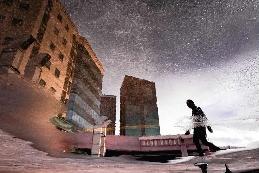 Reflection of city buildings and a walking silhouette distorted in a puddle, creating an abstract and textured street photography scene with muted tones and dynamic composition.