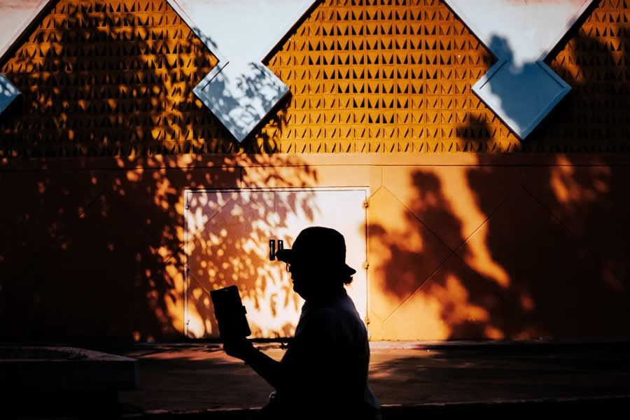 Silhouette of a person reading in front of an orange wall with geometric patterns and tree shadows, creating a balanced composition with strong light and contrast in street photography.