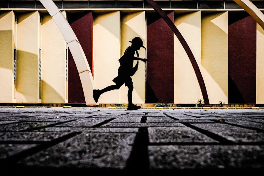 Silhouette of a person walking across a textured pavement with bold geometric architectural background, featuring strong contrast, curved lines, and dramatic shadows in a street photography scene.