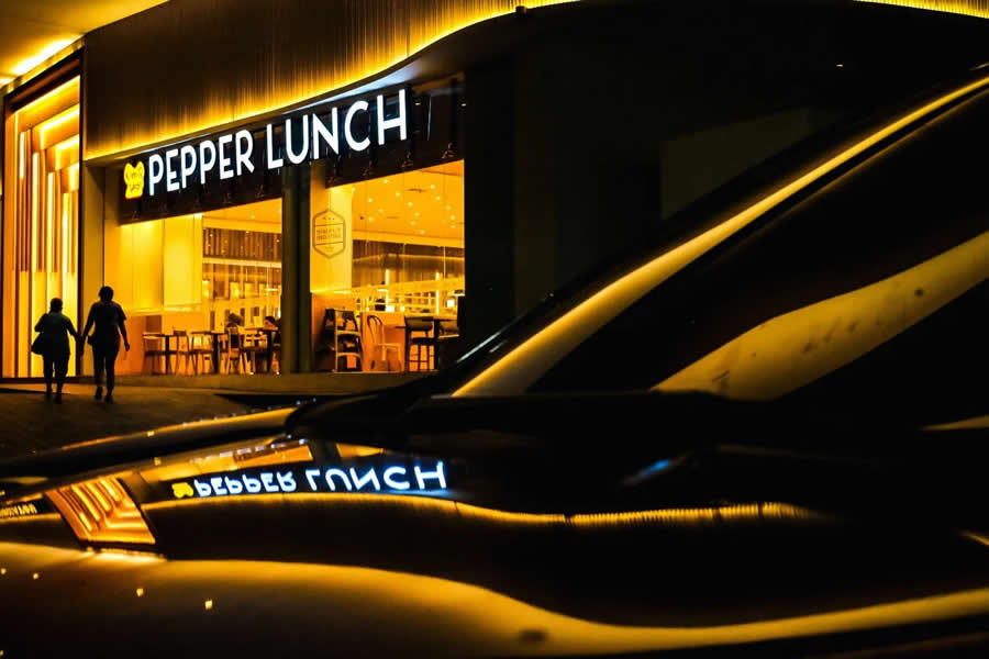 Brightly lit restaurant at night with warm yellow lighting, reflected on a car surface, while two people walk nearby, creating a cinematic street photography scene with strong reflections and contrast.