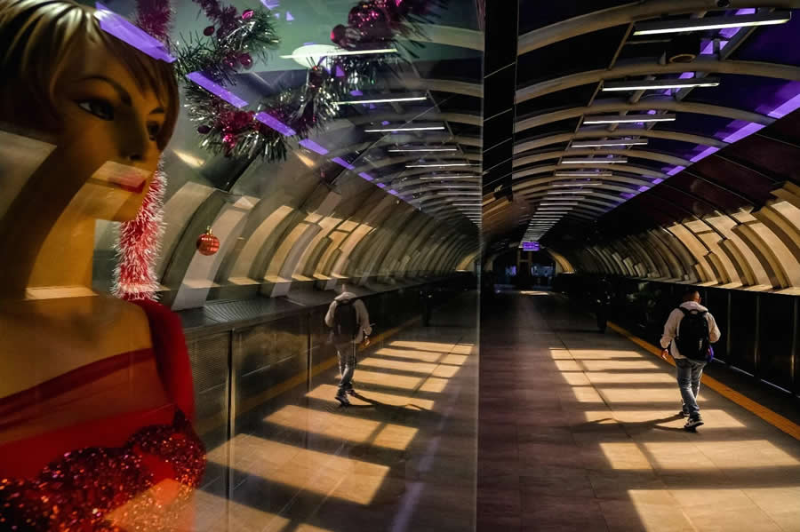 Reflection of a mannequin in a shop window overlapping with a tunnel walkway, where a person walks through repeating architectural lines, creating a layered and surreal street photography composition.