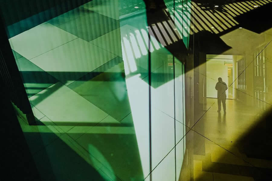 Silhouette of a person seen through layered glass reflections with green and yellow tones, creating geometric shapes and abstract patterns in a modern architectural street photography scene.