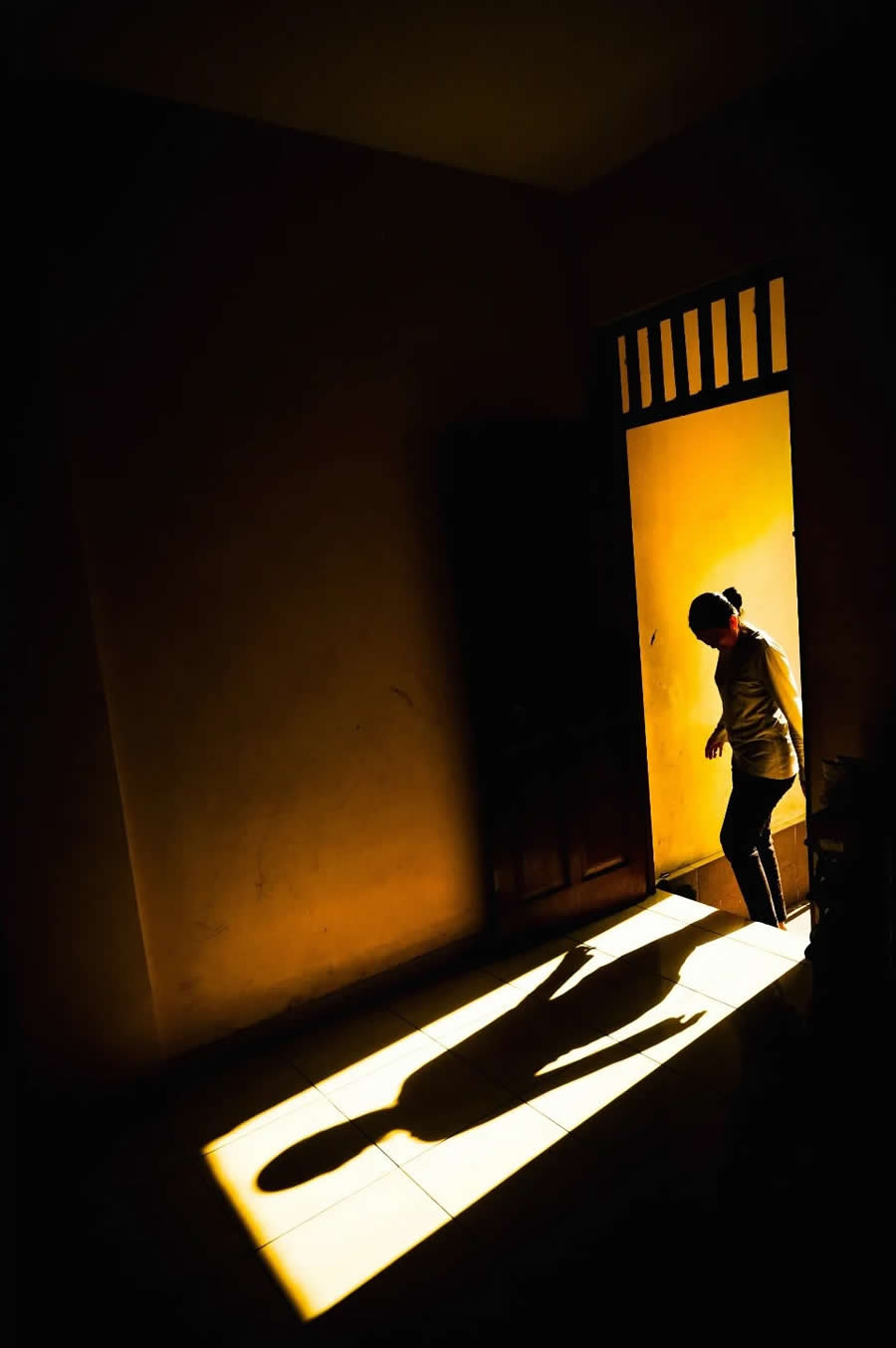 Person stepping through a doorway with warm sunlight casting a long shadow on the floor, creating strong contrast and a minimal, dramatic composition in a street-style photography scene.