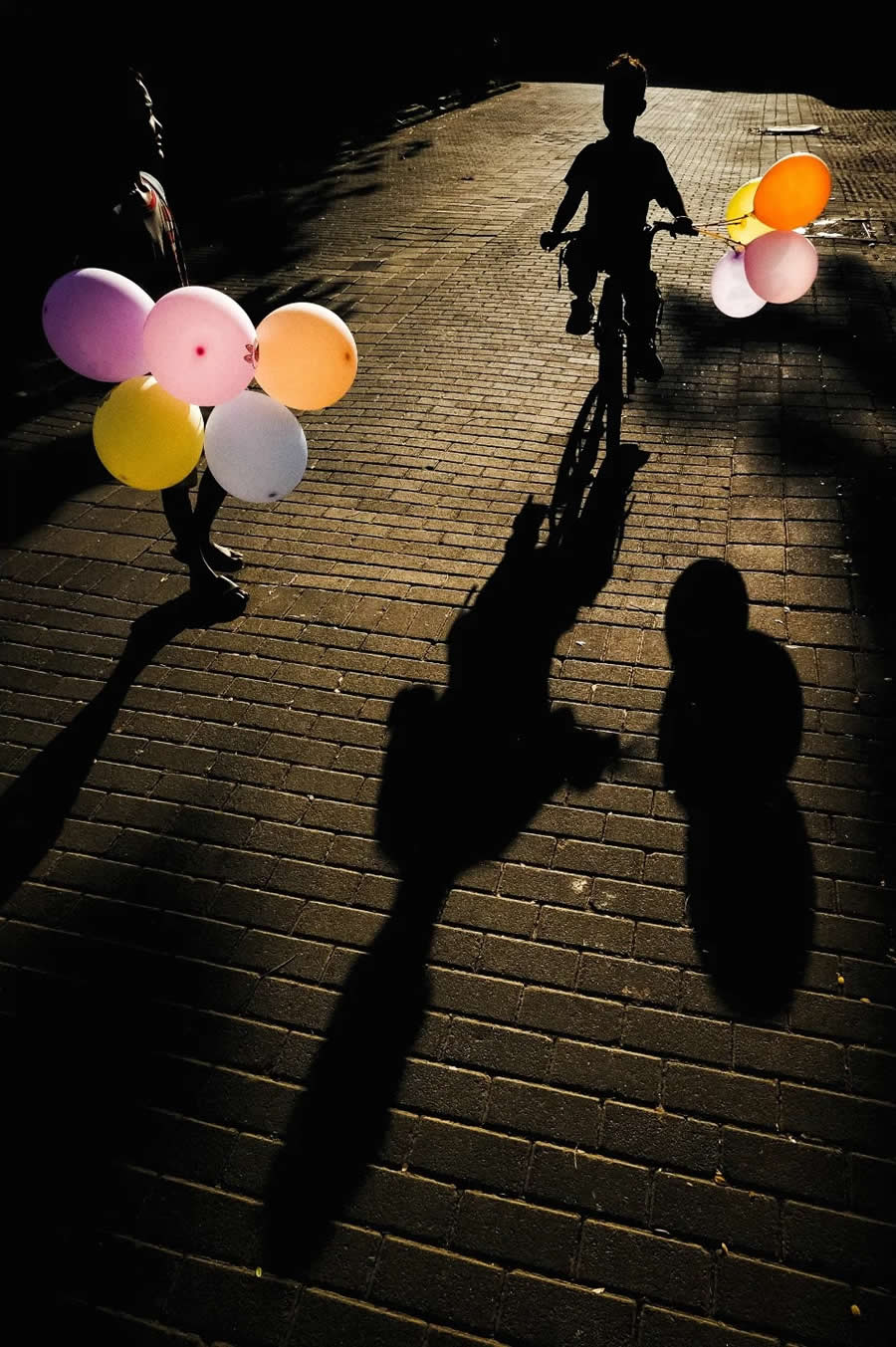 Silhouettes of children walking with colorful balloons, casting long shadows on a sunlit brick pathway, creating strong contrast and a dynamic composition in street photography.