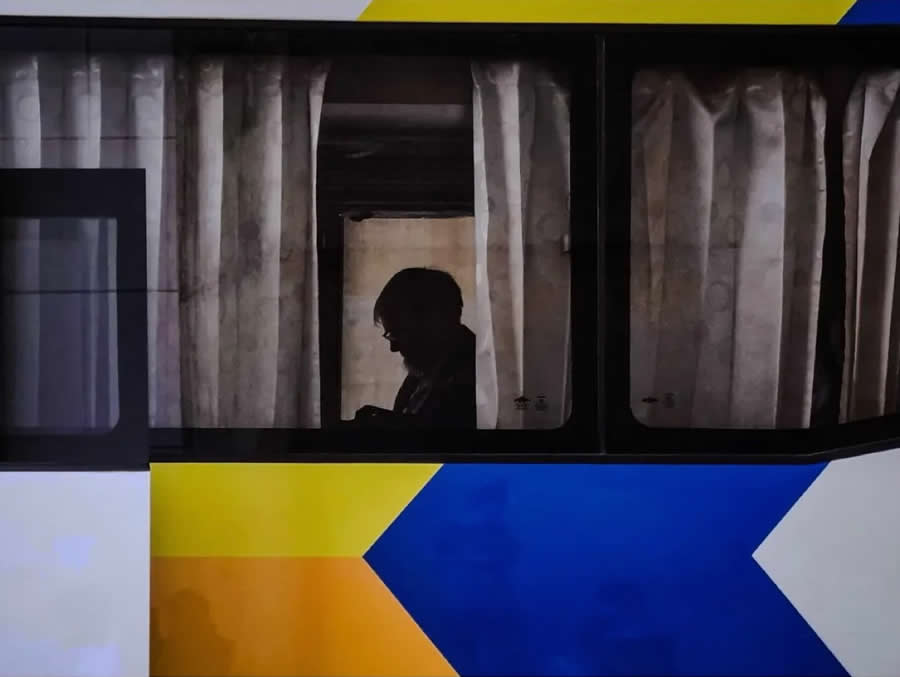 Silhouette of a person inside a bus or train window with curtains, framed by bold geometric blue, yellow, and white patterns, creating a layered and contemplative street photography composition.