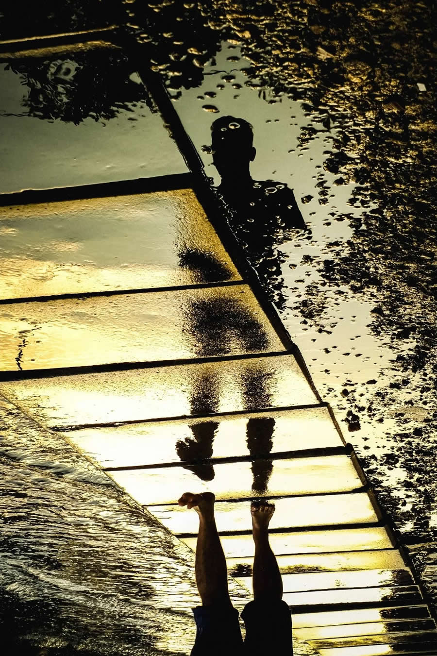 Upside-down reflection of a person in a golden puddle with visible legs and textured water surface, creating an abstract and surreal street photography composition with strong contrast and light.