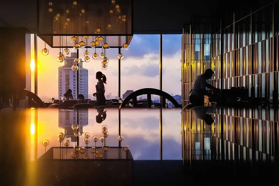 Silhouettes of people inside a modern interior with large windows and sunset light, reflected on a glossy surface with hanging lights, creating a symmetrical and atmospheric street-style photography scene.