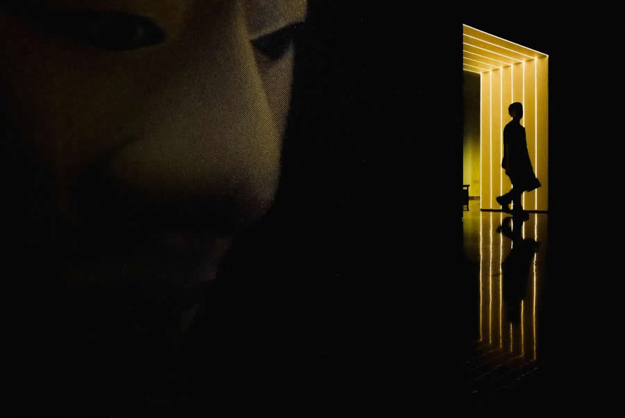 Partial face in deep shadow alongside a brightly lit corridor with a silhouetted person walking, reflected on the floor, creating a high-contrast, cinematic street photography composition.