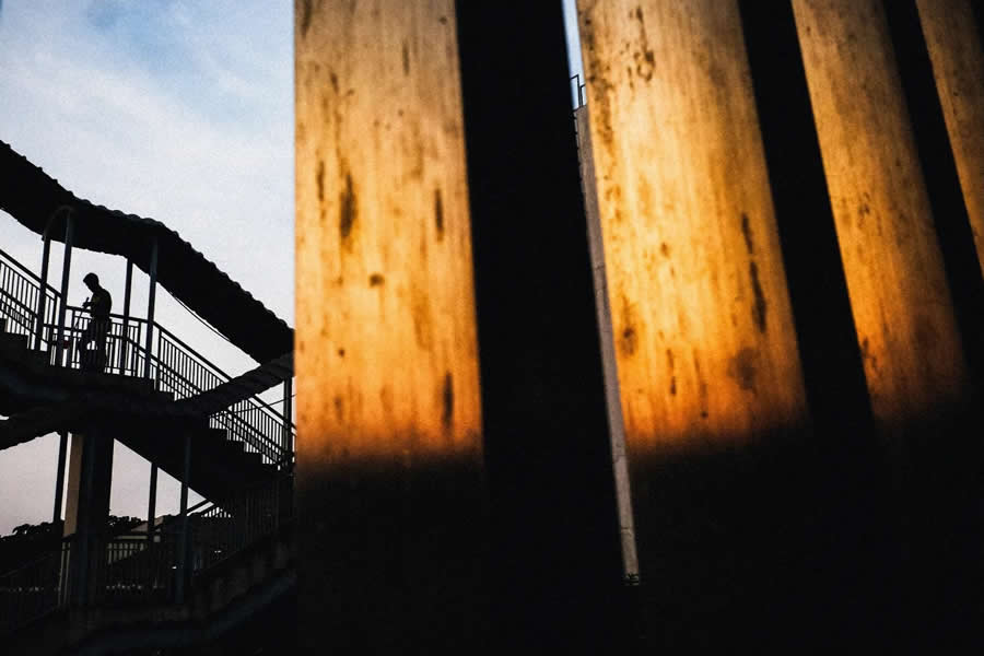 Silhouette of a person walking on a staircase in the background, contrasted with glowing textured panels in the foreground, creating a layered composition with strong light and shadow in street photography.