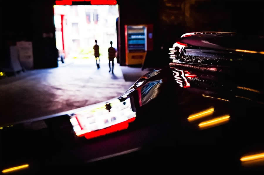 Reflections of neon lights on a car surface at night with two silhouetted figures walking in the background, creating a layered and cinematic street photography composition with strong contrast and color.