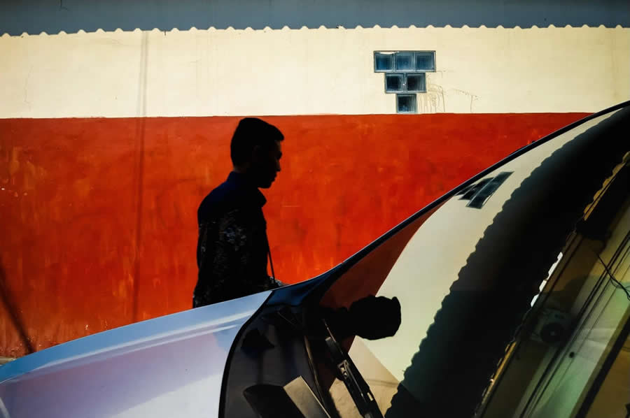Silhouette of a man walking past a red and white wall with a car in the foreground reflecting his figure, creating strong contrast and clean geometric composition in street photography.
