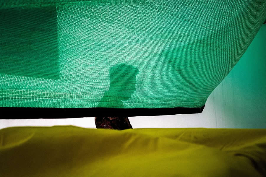 Silhouette of a person seen through a green translucent fabric with a contrasting yellow foreground, creating a minimal, layered composition with soft light and abstract textures in street photography.
