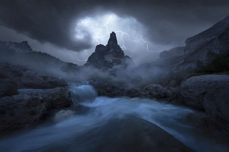 Lightning striking behind a rocky mountain peak with mist, flowing water, and dark storm clouds creating a dramatic landscape.