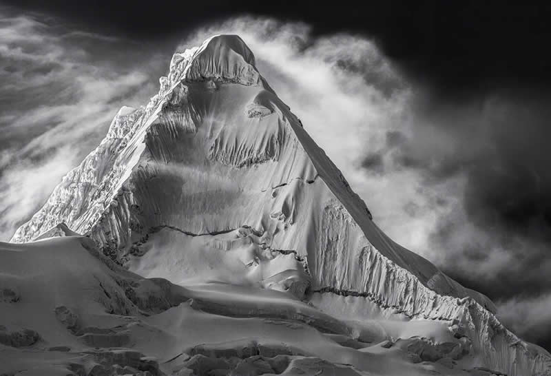 Black-and-white image of Alpamayo mountain showing sharp ridges, steep snowy slopes, and dramatic cloud formations.
