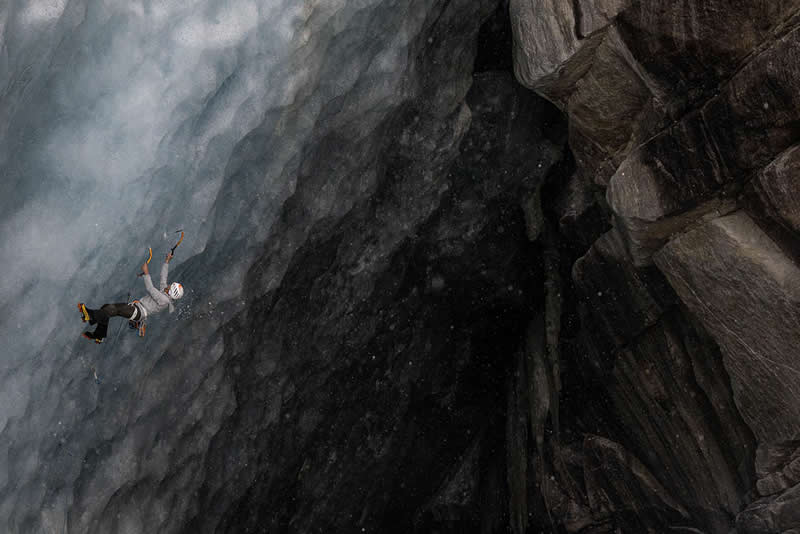 A climber falling on a steep icy wall beside a dark rocky cliff, captured mid-air in a dramatic alpine scene.