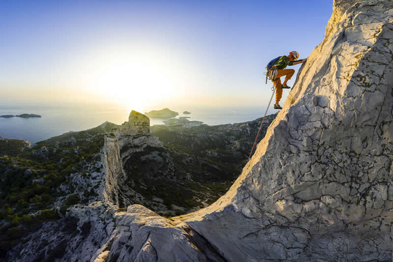 A climber scaling a steep rocky ridge above a coastal landscape at sunrise, with ocean and islands visible in the background.