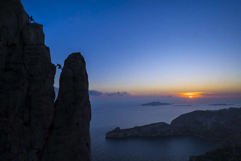 Silhouetted climbers on tall sea cliffs at sunset overlooking the ocean and distant islands under a colorful sky.