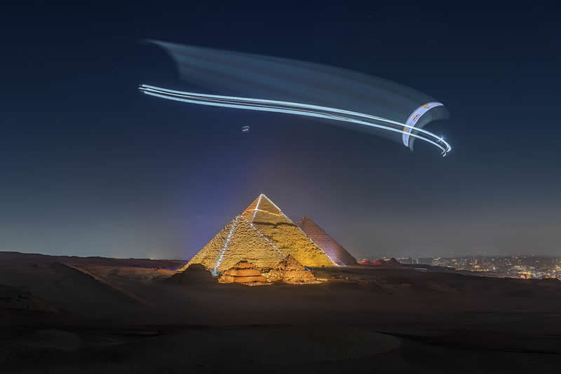 Paraglider flying at night above illuminated pyramids, leaving light trails in the sky over a desert landscape.