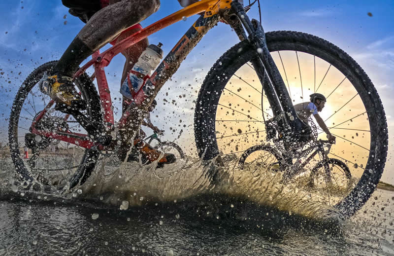Close-up of cyclists riding through water and mud, with splashes frozen mid-air and wheels in motion during a race.