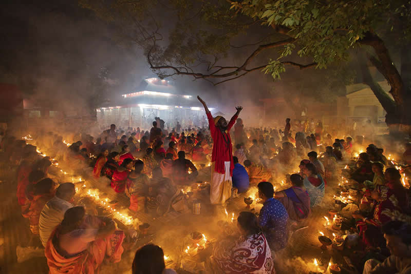 Crowd of people performing a ritual with candles and fire at night, with one person standing in the center raising their arms.