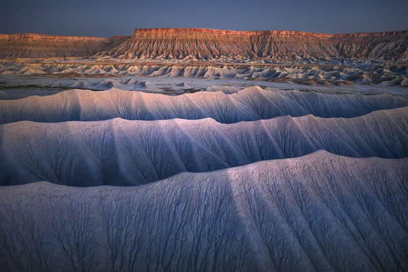 Eroded badlands with wave-like ridges in the foreground and a sunlit cliff in the background under soft evening light.