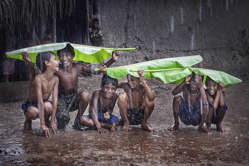 Children playing in heavy rain, using banana leaves as umbrellas, smiling and crouching together in a muddy rural setting.