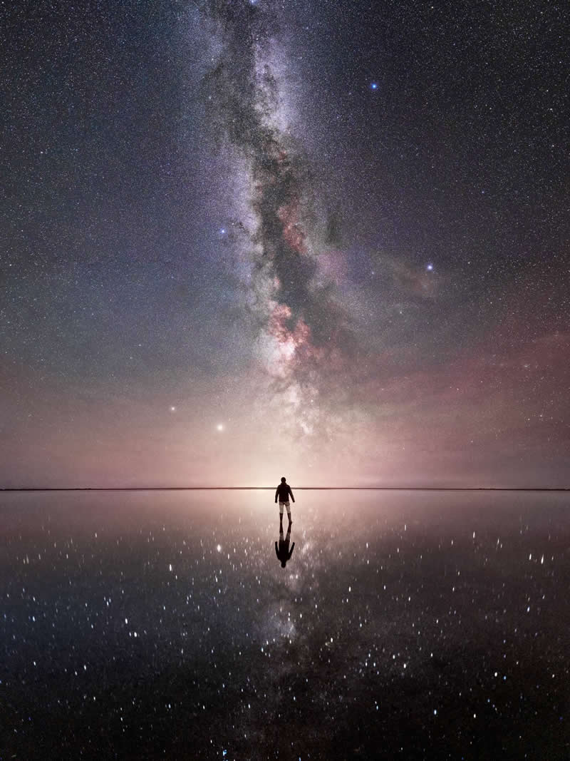 Beyond the Stars: How Mads Peter Iversen Captures the Milky Way Like No One Else 27 A person’s silhouette stands on a glossy, reflective surface under a brilliant Milky Way. The stars and swirling colors in the sky are mirrored perfectly below, turning the night into a surreal, otherworldly scene.