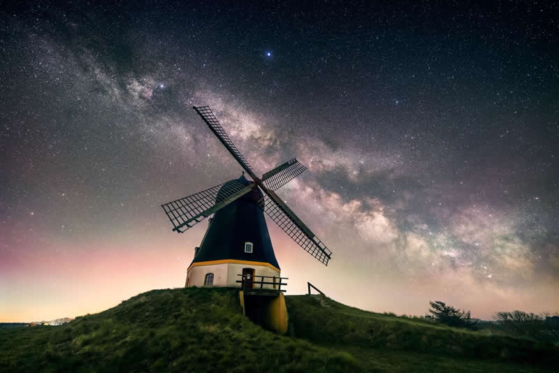 Beyond the Stars: How Mads Peter Iversen Captures the Milky Way Like No One Else 31 Traditional windmill on a grassy hill under a glowing Milky Way galaxy with stars stretching across the night sky.