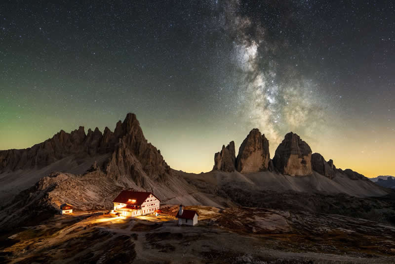 Beyond the Stars: How Mads Peter Iversen Captures the Milky Way Like No One Else 33 A glowing mountain cabin sits below dramatic rocky peaks, while the Milky Way arcs across a crisp, clear night sky above.