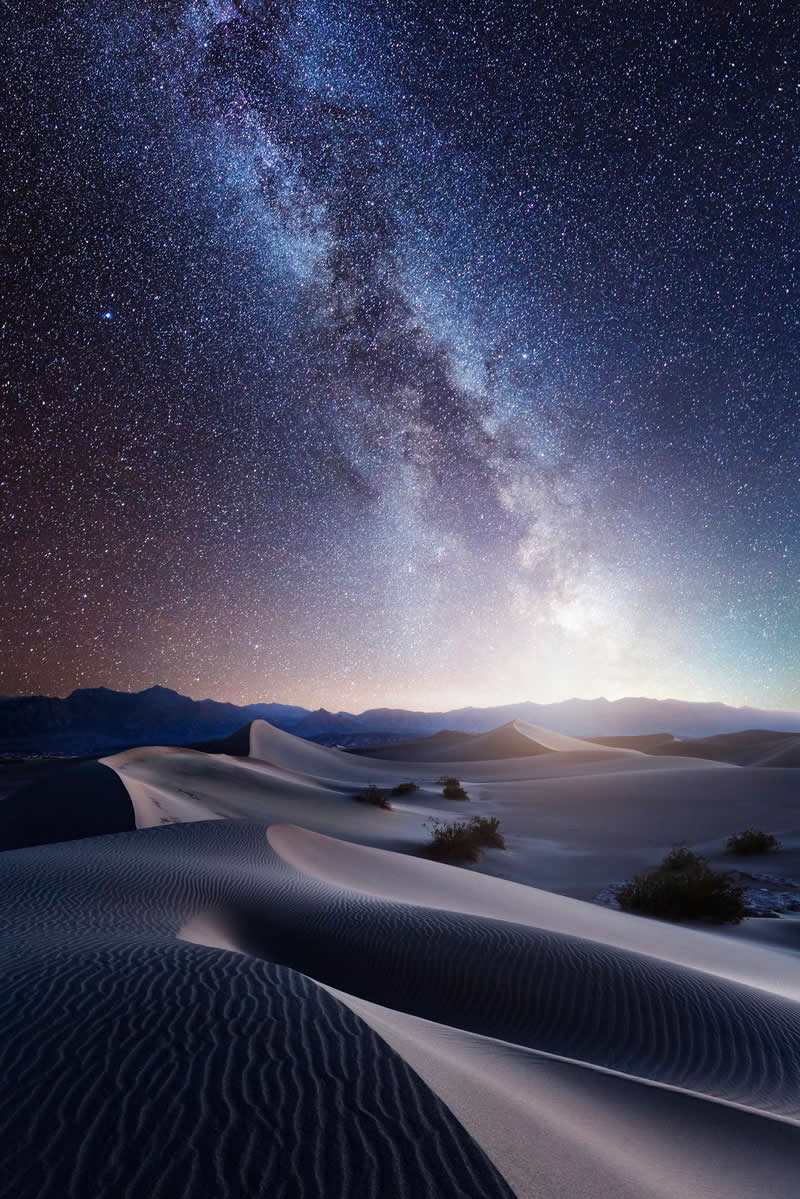 Beyond the Stars: How Mads Peter Iversen Captures the Milky Way Like No One Else 41 Milky Way galaxy shining above smooth desert sand dunes with mountains in the distance under a clear star-filled night sky.