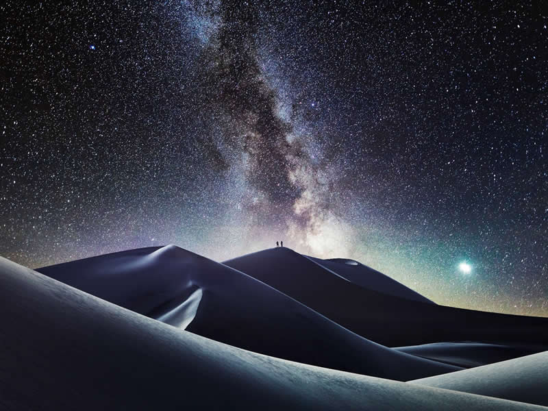 Beyond the Stars: How Mads Peter Iversen Captures the Milky Way Like No One Else 42 Two people standing on sand dunes under a bright Milky Way galaxy with countless stars filling the night sky above the desert landscape.