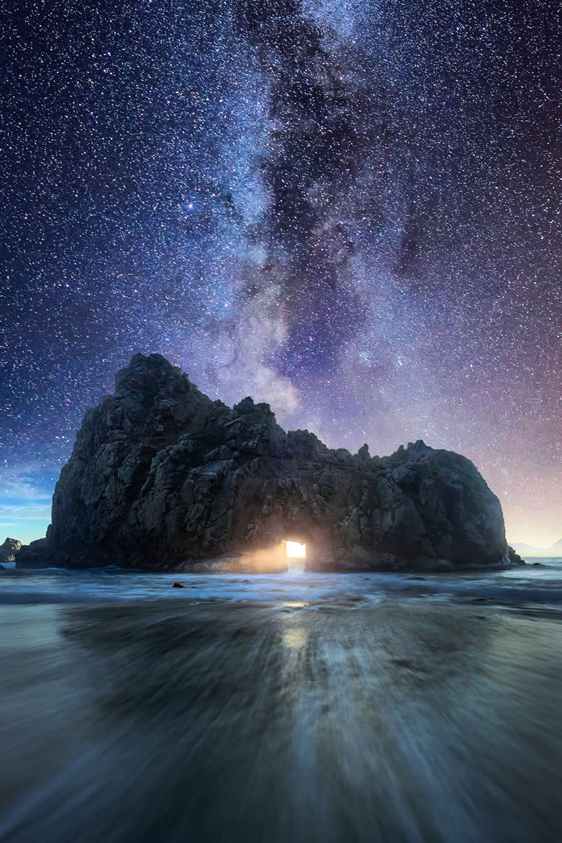 Beyond the Stars: How Mads Peter Iversen Captures the Milky Way Like No One Else 44 Milky Way galaxy rising above a rocky sea arch with light glowing through the opening while waves blur across the shoreline at night.