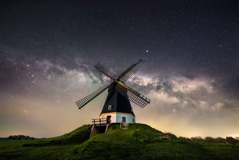Beyond the Stars: How Mads Peter Iversen Captures the Milky Way Like No One Else 45 Traditional windmill on a grassy hill beneath the Milky Way galaxy stretching across a clear star-filled night sky.