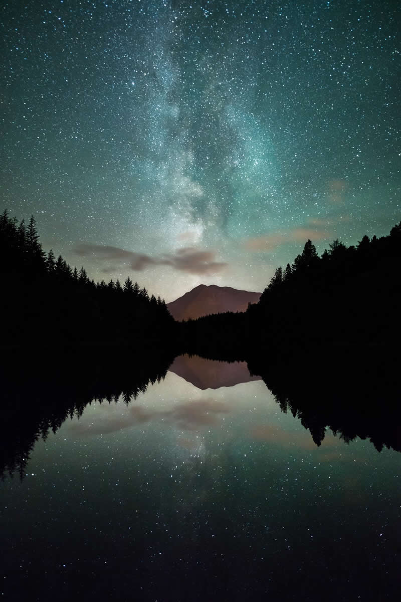 Beyond the Stars: How Mads Peter Iversen Captures the Milky Way Like No One Else 49 Milky Way galaxy reflected in a calm mountain lake surrounded by dark forest silhouettes with a distant mountain under a star-filled night sky.