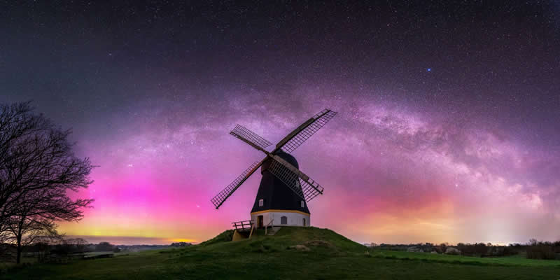 Beyond the Stars: How Mads Peter Iversen Captures the Milky Way Like No One Else 50 Windmill on a grassy hill under a colorful Milky Way arch with pink and purple night sky glowing above the landscape.