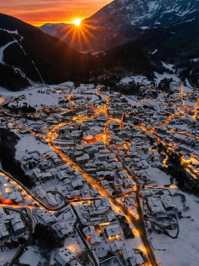 Aerial sunset view of a snow-covered alpine village with glowing golden streetlights and dramatic sun rays over mountains during winter twilight.
