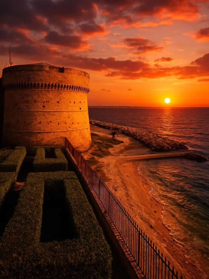 Historic coastal fortress overlooking the sea during a dramatic orange sunset with glowing clouds, warm light on stone walls, and calm ocean waves along the shoreline.