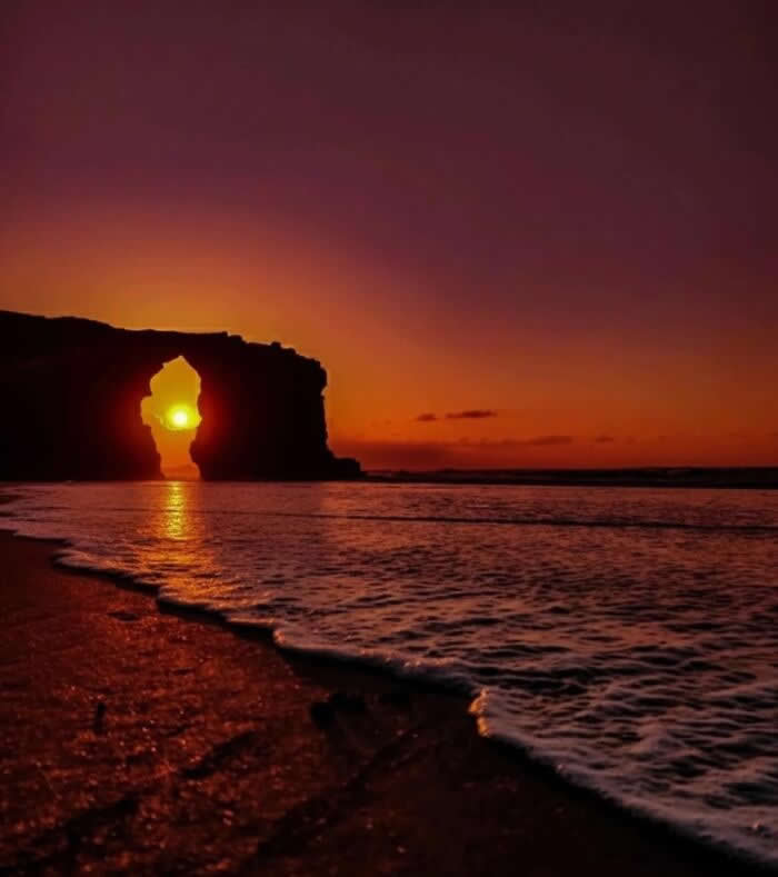 Sunset shining through a natural sea arch rock formation on a beach with waves and deep orange sky reflecting on the ocean at twilight.