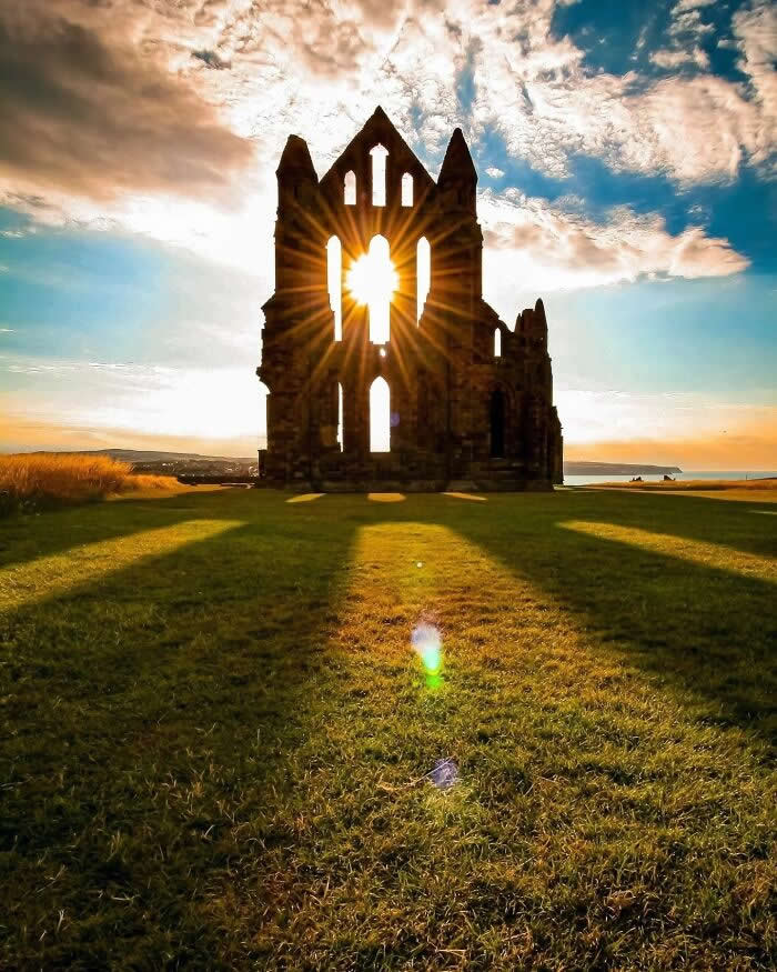 Sunburst shining through the windows of an ancient abbey ruin at sunset with long shadows across green grass and dramatic clouds in the sky.