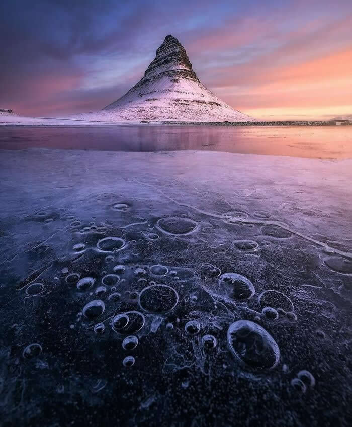 Snow-covered mountain reflected on frozen lake with trapped ice bubbles in foreground under a soft pastel sunset sky creating dramatic winter landscape scenery.