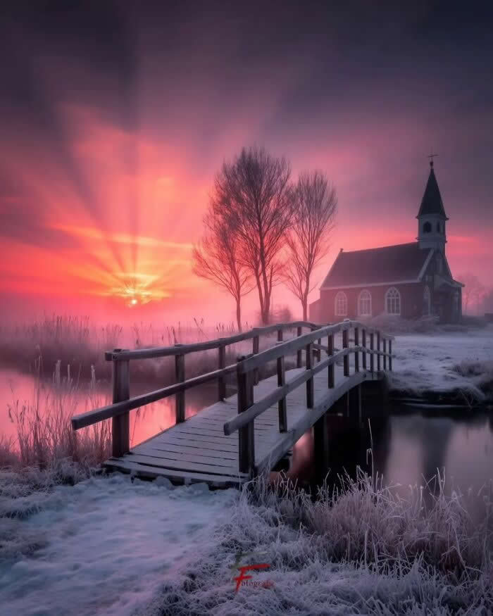 Wooden bridge over frosty pond leading to a small country church during a pink and golden winter sunset with misty sky and glowing sun rays.